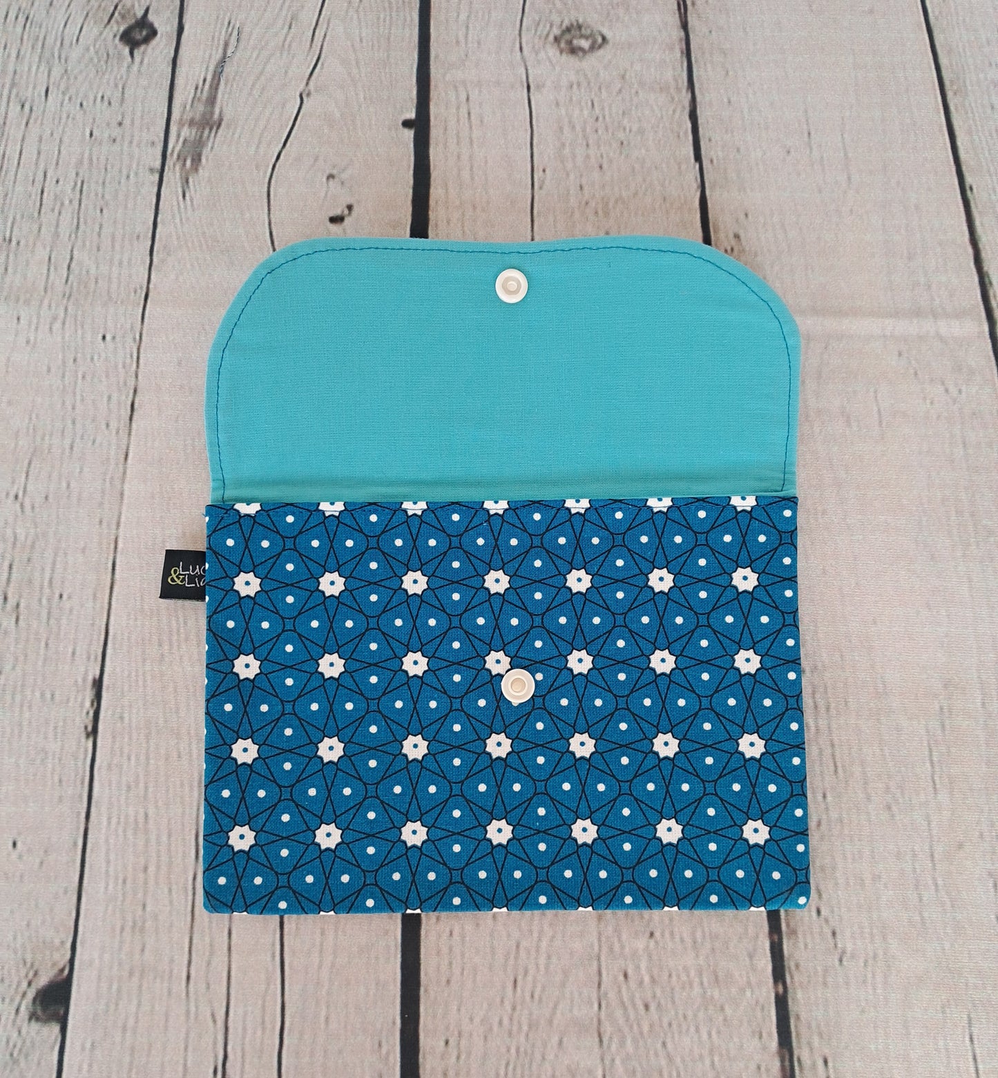 Estuche multiusos azulon: Handcrafted multipurpose clutch in turquoise and navy blue with white daisy pattern, displayed open on whitewashed wooden surface. Features snap closure and dual-tone design with solid turquoise interior and floral print exterior.