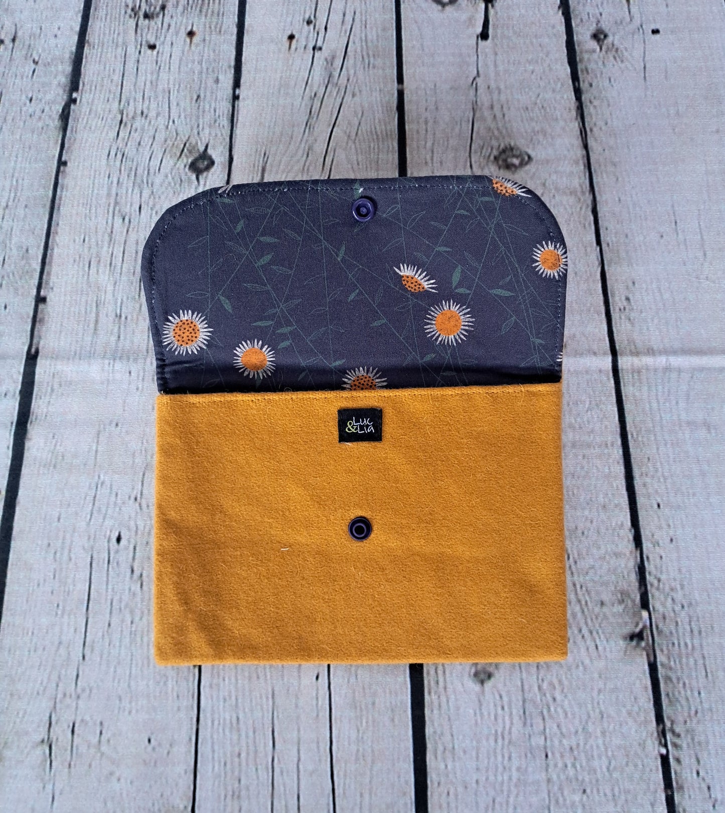 Estuche multiusos: Handcrafted multipurpose case featuring a navy blue fabric with delicate sunflower pattern and mustard yellow suede flap, displayed on weathered white wooden background. Sustainable accessory for documents and personal items.