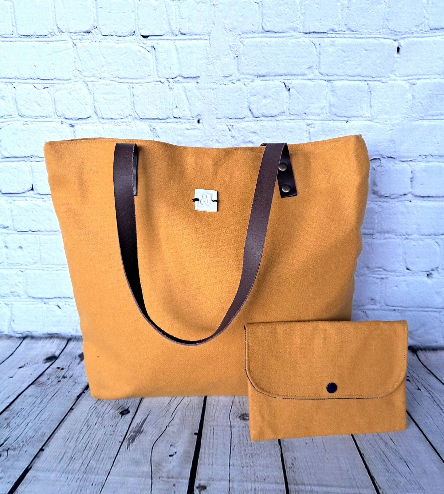 Conjunto Provence bolso y estuche - Handcrafted mustard yellow canvas tote bag with brown leather straps and matching clutch pouch, displayed against white brick wall on wooden floor. Sustainable fashion accessories set made from natural materials.