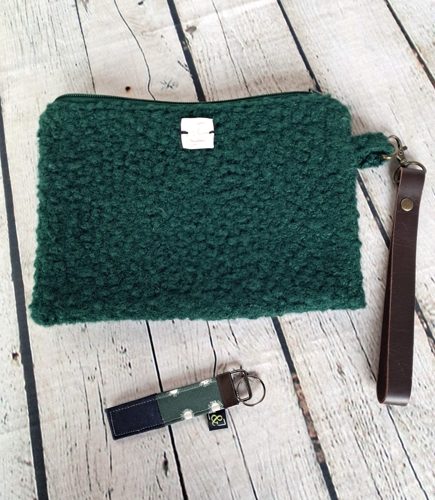 Conjunto bolso y llavero: Handcrafted emerald green textured clutch with brown leather wrist strap and matching keychain, photographed on whitewashed wooden planks. Sustainable fashion accessory set showcasing artisanal craftsmanship and eco-friendly materials.