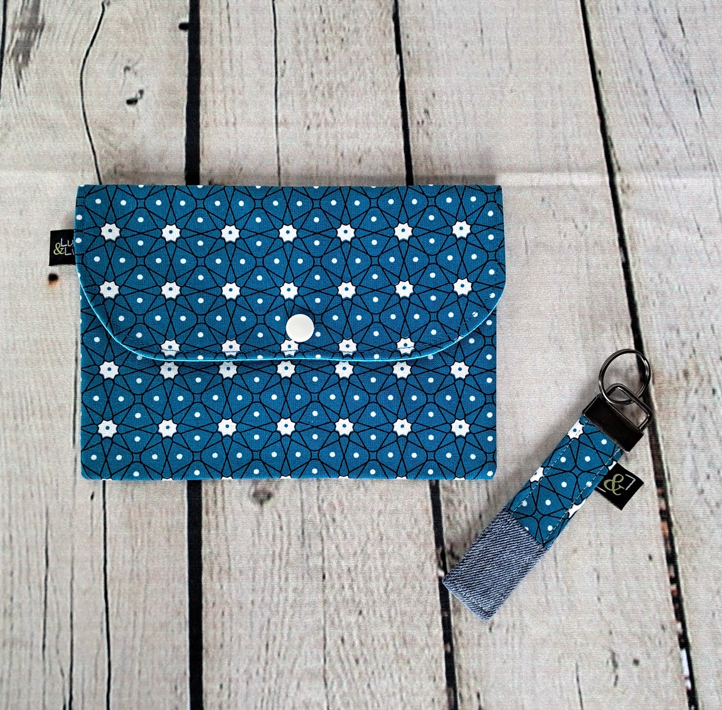 Estuche y llavero handmade case and keychain set featuring blue fabric with white floral geometric pattern, displayed on whitewashed wooden planks. Eco-friendly accessory crafted from sustainable materials with snap button closure and coordinating fabric key fob.