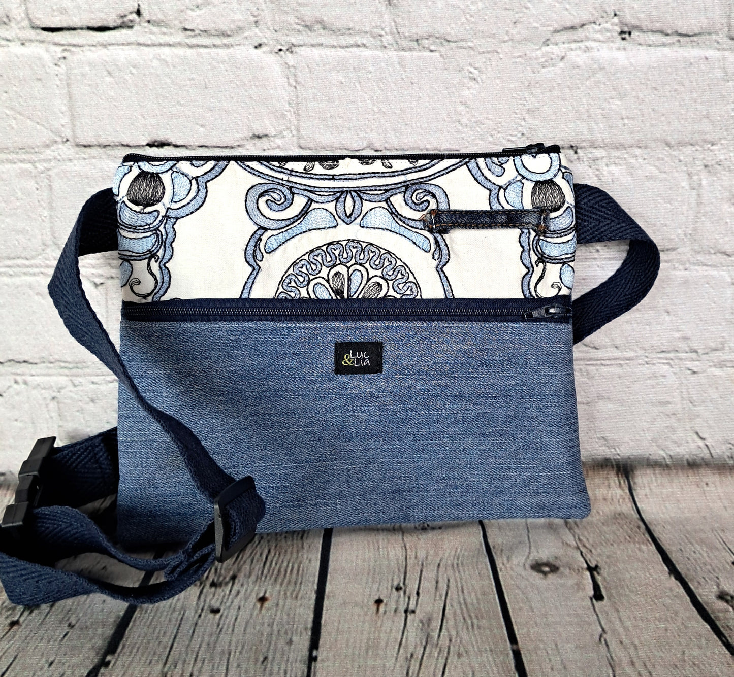 Denim and embroidered blue fanny pack (Rinonera diseno denim y bordado azul) featuring white fabric with blue ornate floral pattern, adjustable navy strap, and convenient zipper closure, displayed on wooden surface against white brick wall.
