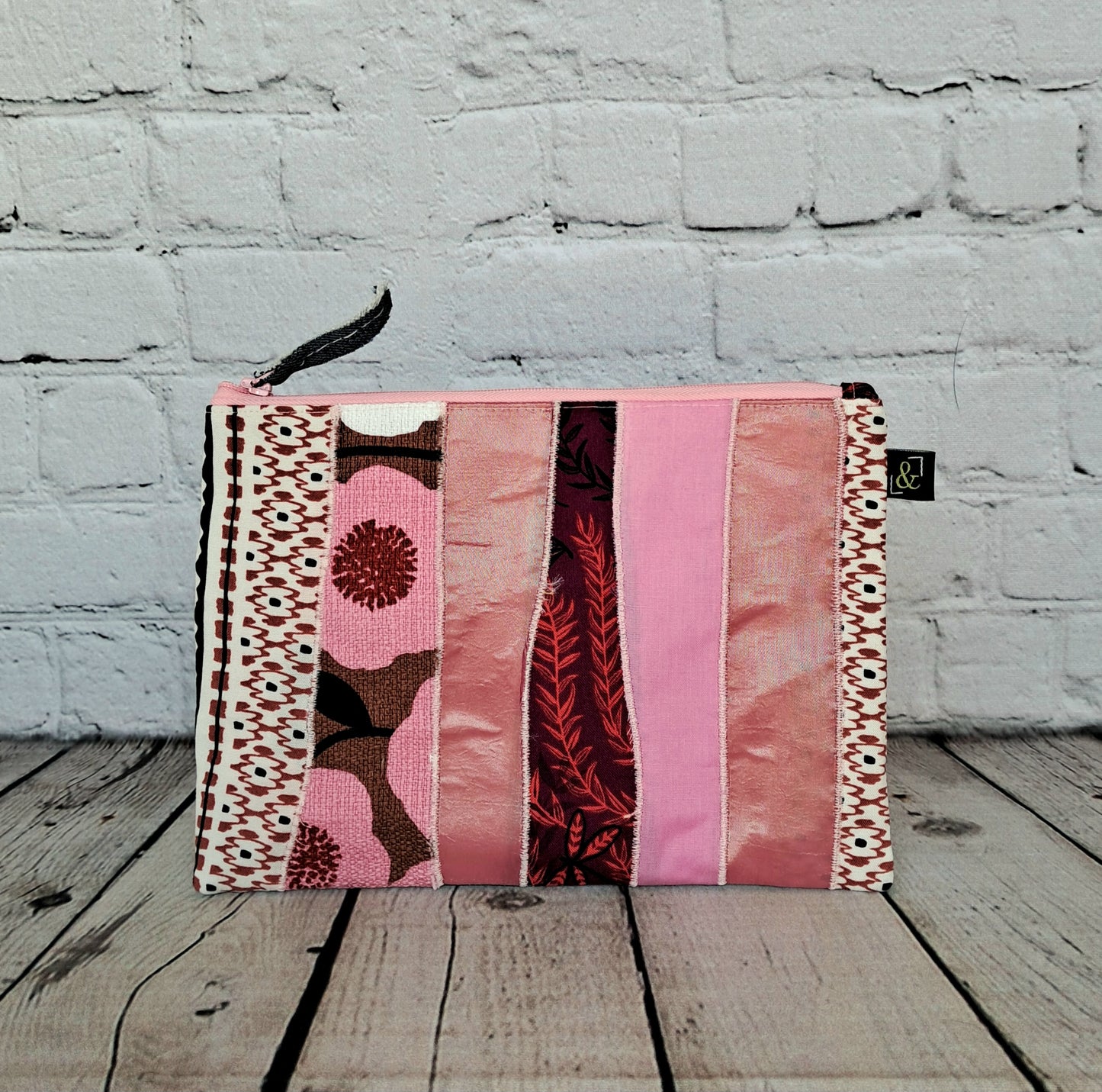 Estuche cremallera de retales rosas - Handmade patchwork pencil case featuring pink fabric strips in various patterns and textures, including floral prints and geometric designs, with black zipper closure against white brick backdrop, showcasing sustainable craftsmanship and unique design