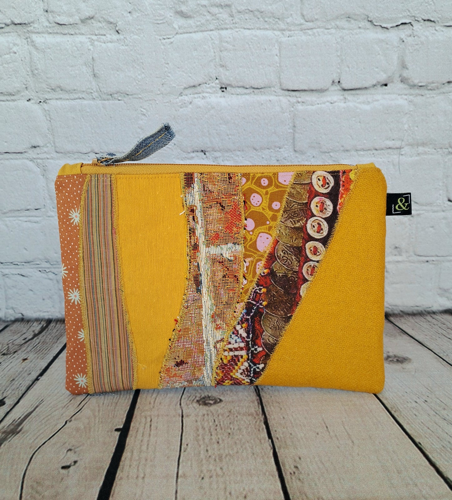 Yellow patchwork zipper pouch, Estuche cremallera de retales amarillos, handmade pencil case featuring unique fabric combinations in yellow, orange and brown tones, displayed against white brick wall background. Eco-friendly upcycled textile design with decorative pattern strips and gray zipper.