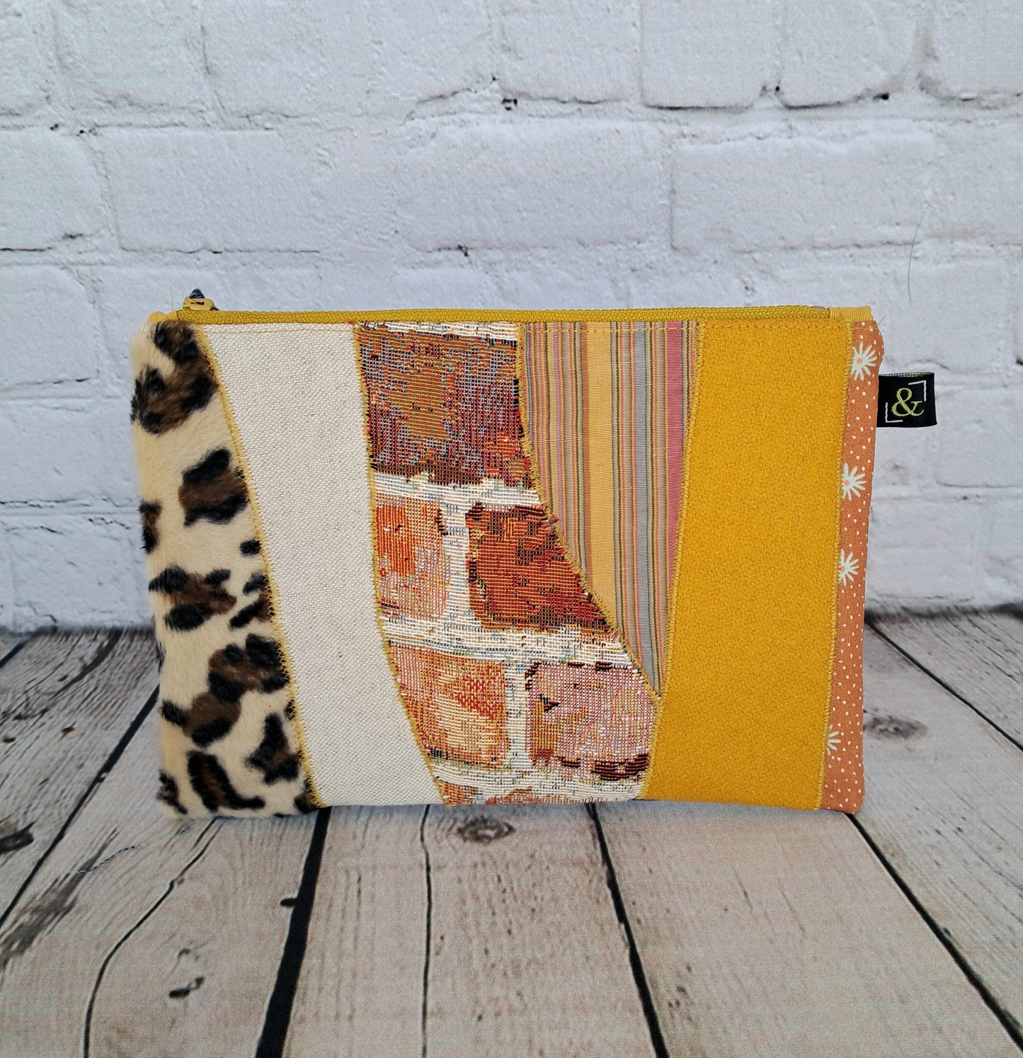 Handmade yellow patchwork zipper pouch 'Estuche cremallera de retales amarillos' featuring unique fabric combinations including animal print, striped patterns, and solid yellow sections, displayed on white brick background. Sustainable accessory measures 22 x 16 cm.