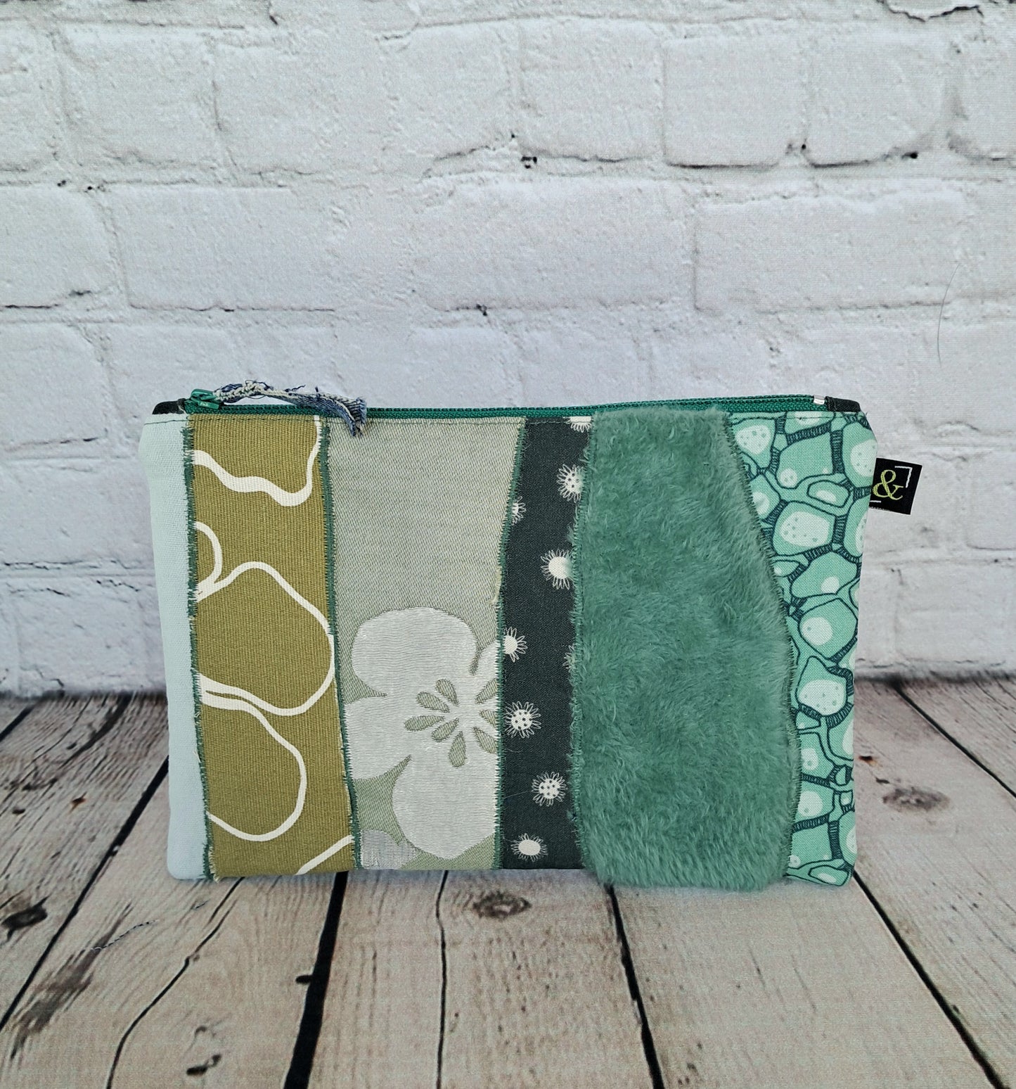 Estuche cremallera de retales verdes - Handcrafted zippered pouch featuring patchwork design in various shades of green and white, combining floral patterns, textures, and geometric prints, set against rustic wooden floor and white brick wall background.