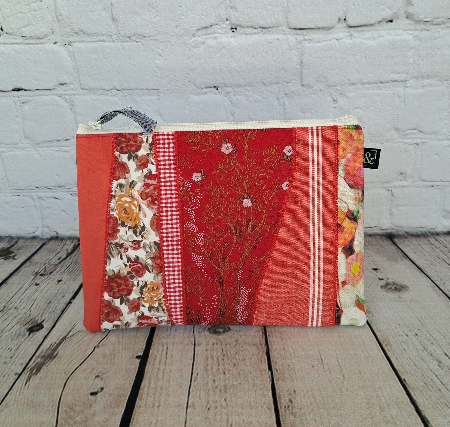 Handmade zipper case Estuche cremallera de retales rojos featuring patchwork design in red tones with floral prints, gingham checks, and decorative patterns. Eco-friendly pencil case made from upcycled fabrics against white brick wall background on wooden surface. Size 22 x 16 cm.