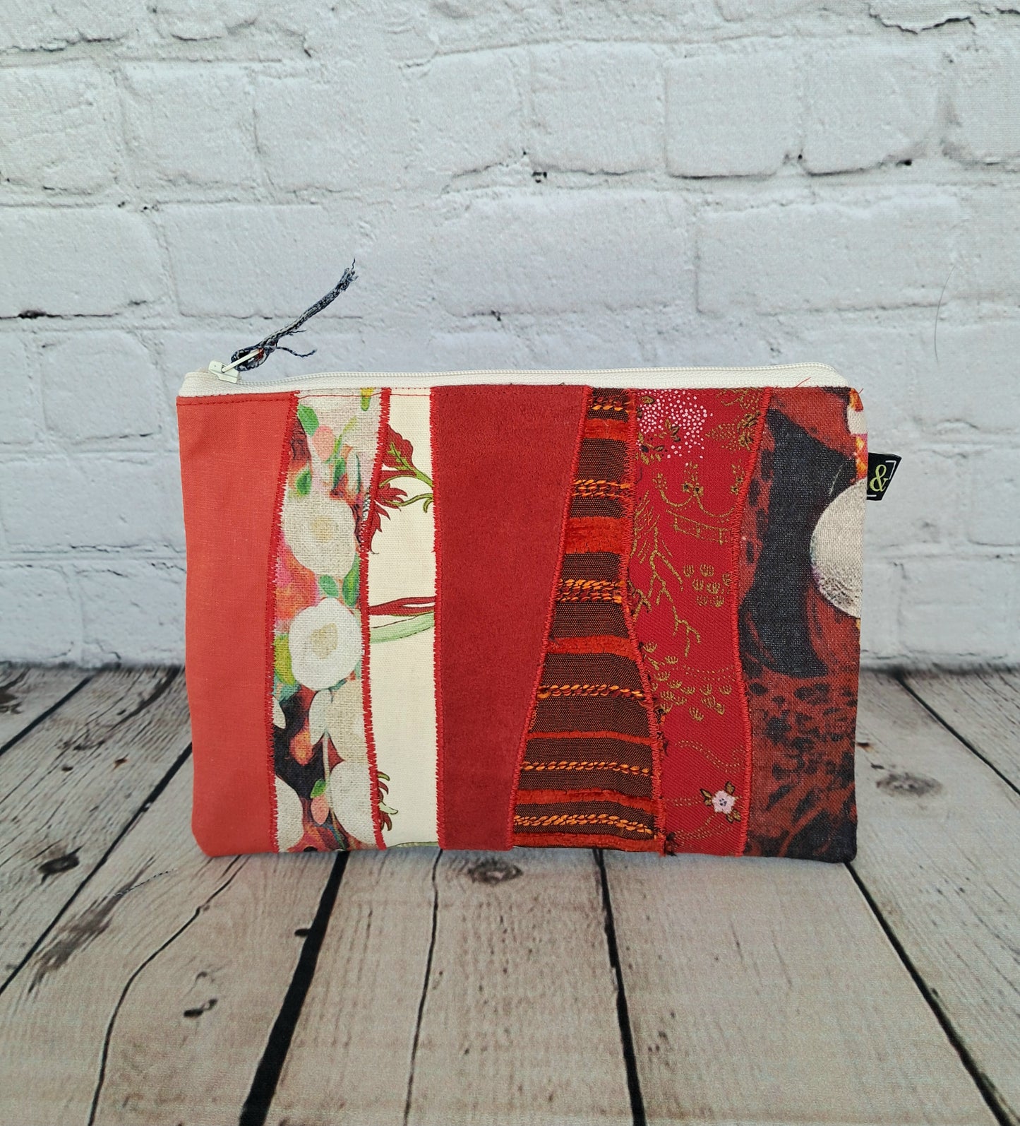 Estuche cremallera de retales rojos - Handmade zipper pouch featuring a patchwork design in various shades of red fabric, combining floral prints and geometric patterns. Displayed on wooden planks against a white brick wall background, showcasing sustainable craftsmanship and unique textile combinations.