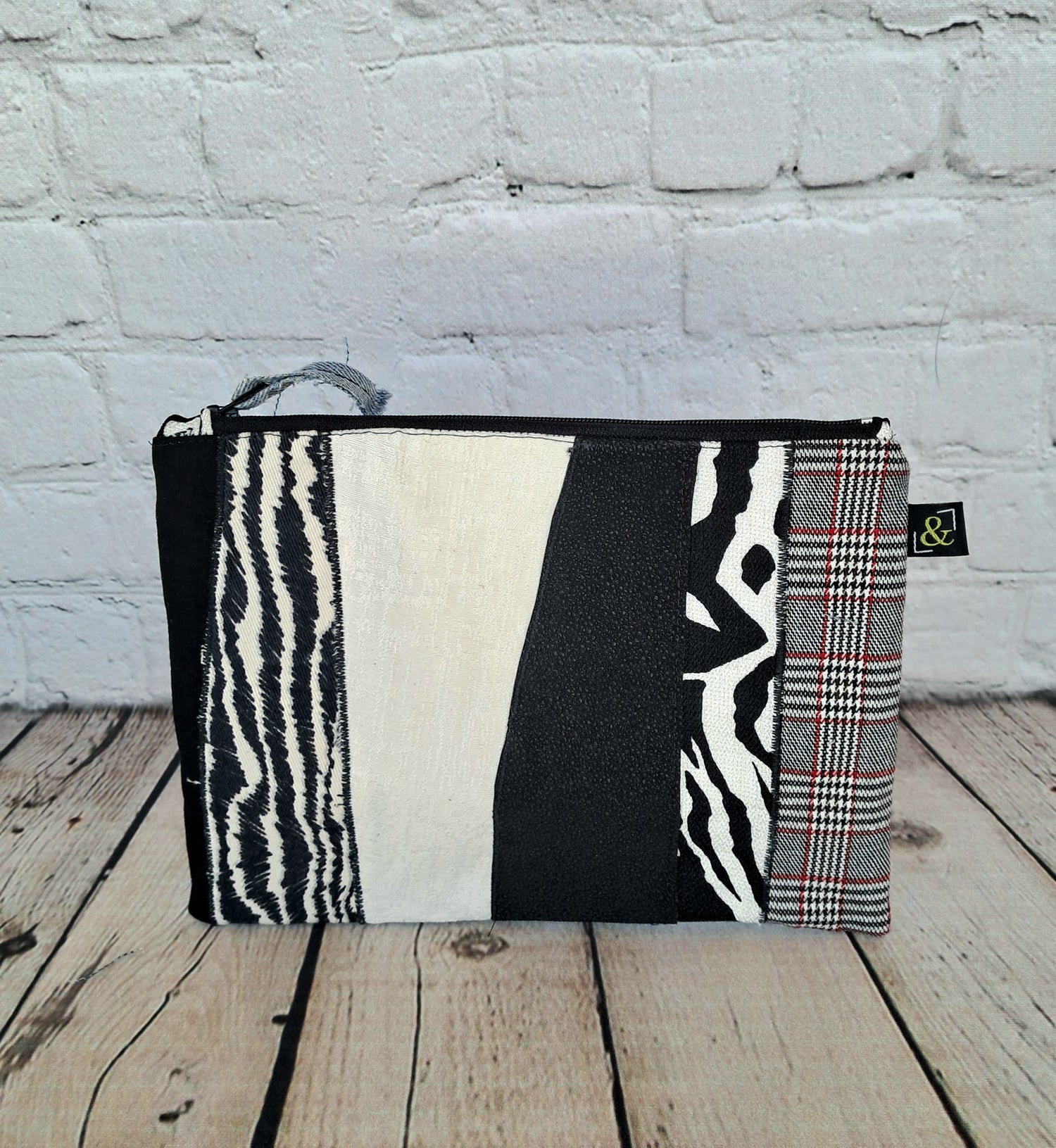 Estuche cremallera de retales blanco y negro - Handcrafted zipper pouch featuring unique patchwork design in black and white patterns, including zebra print, solid blocks, and plaid fabric, displayed on white brick background with wooden floor. Size 22 x 16 cm, showcasing sustainable upcycled materials.