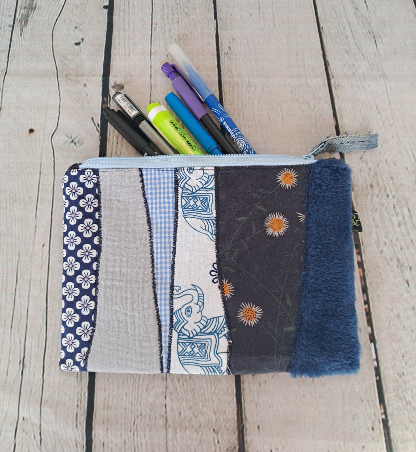 Handmade Blue Patchwork Pencil Case 'Estuche cremallera de retales azules' featuring mixed fabric patterns including floral prints, gingham, and textured materials, with blue zipper closure, shown with markers inside on wooden background. Size 22 x 16 cm.