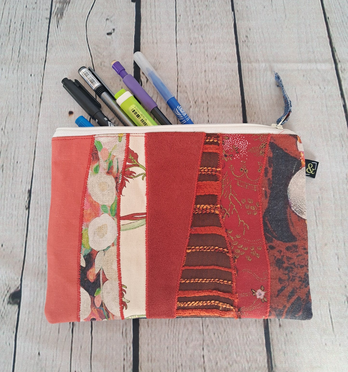 Estuche cremallera de retales rojos - Handcrafted zippered pencil case made from upcycled red fabric patches displaying various patterns and textures, shown filled with pens on whitewashed wooden surface. Sustainable and eco-friendly accessory measuring 22 x 16 cm.