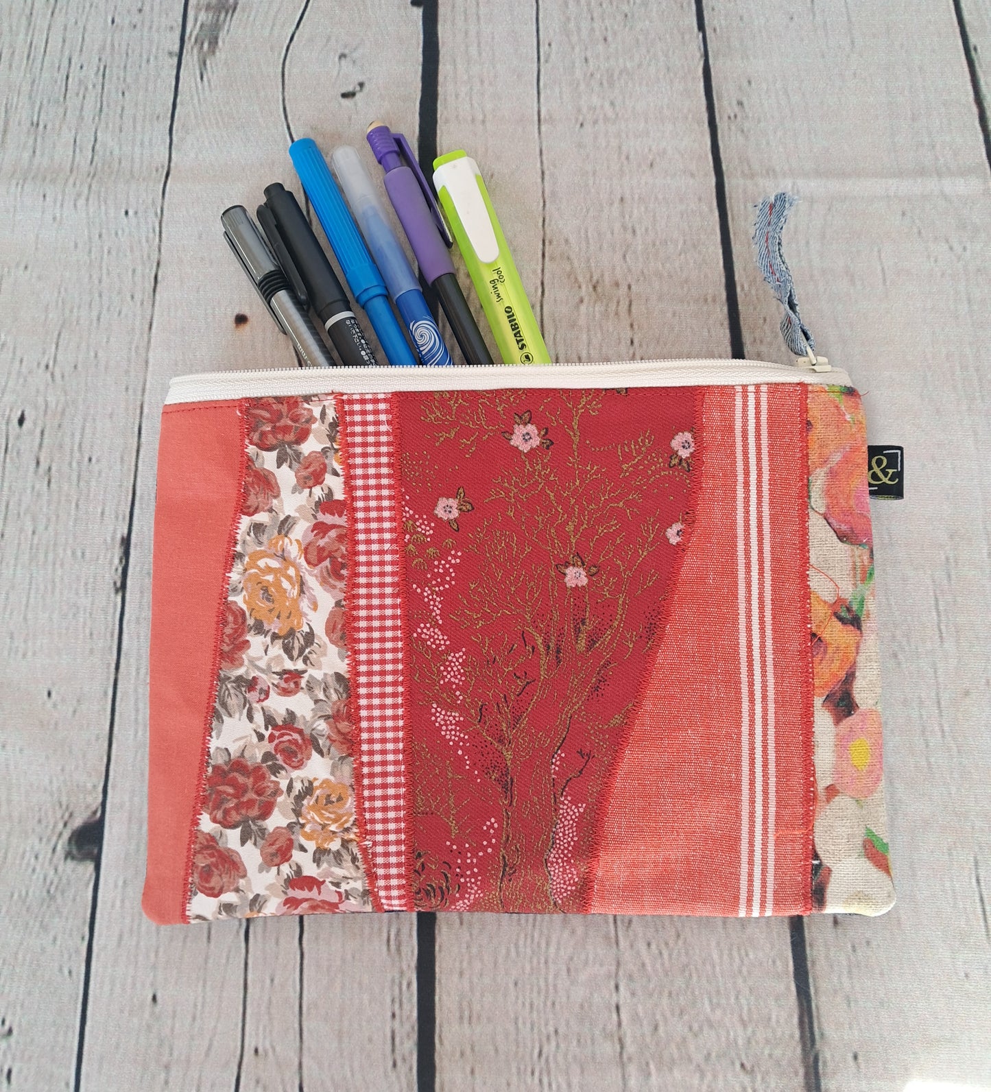 Handmade red patchwork pencil case 'Estuche cremallera de retales rojos' featuring floral patterns, gingham checks, and vintage fabrics, filled with colorful pens on whitewashed wooden background. Sustainable and unique zipper pouch measuring 22 x 16 cm.