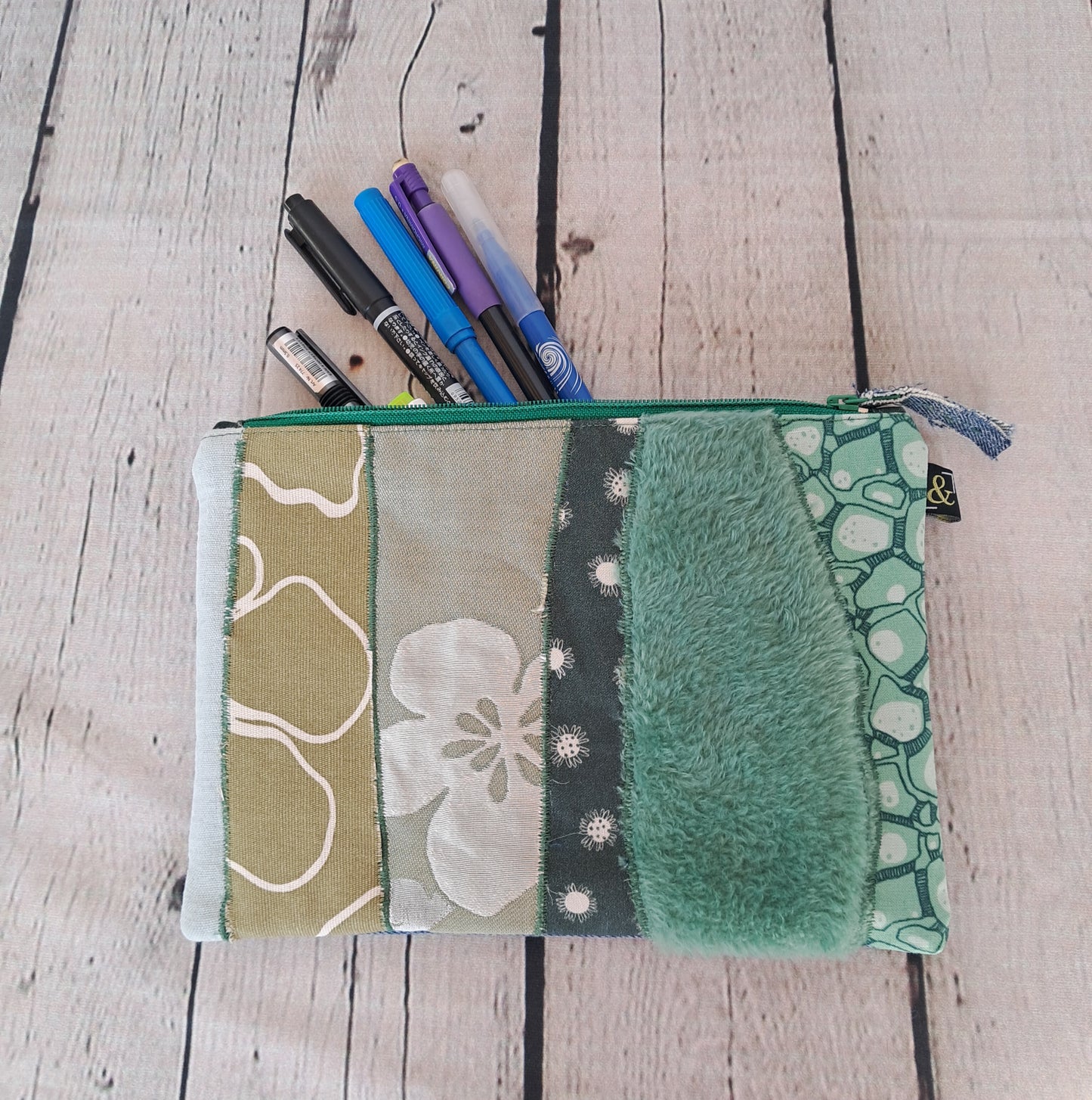 Estuche cremallera de retales verdes: Handmade pencil case featuring patchwork design in green tones with various textures and patterns, shown with pens inside, displayed on white wooden surface. Zipper closure, combining floral prints, fuzzy fabric, and geometric patterns.