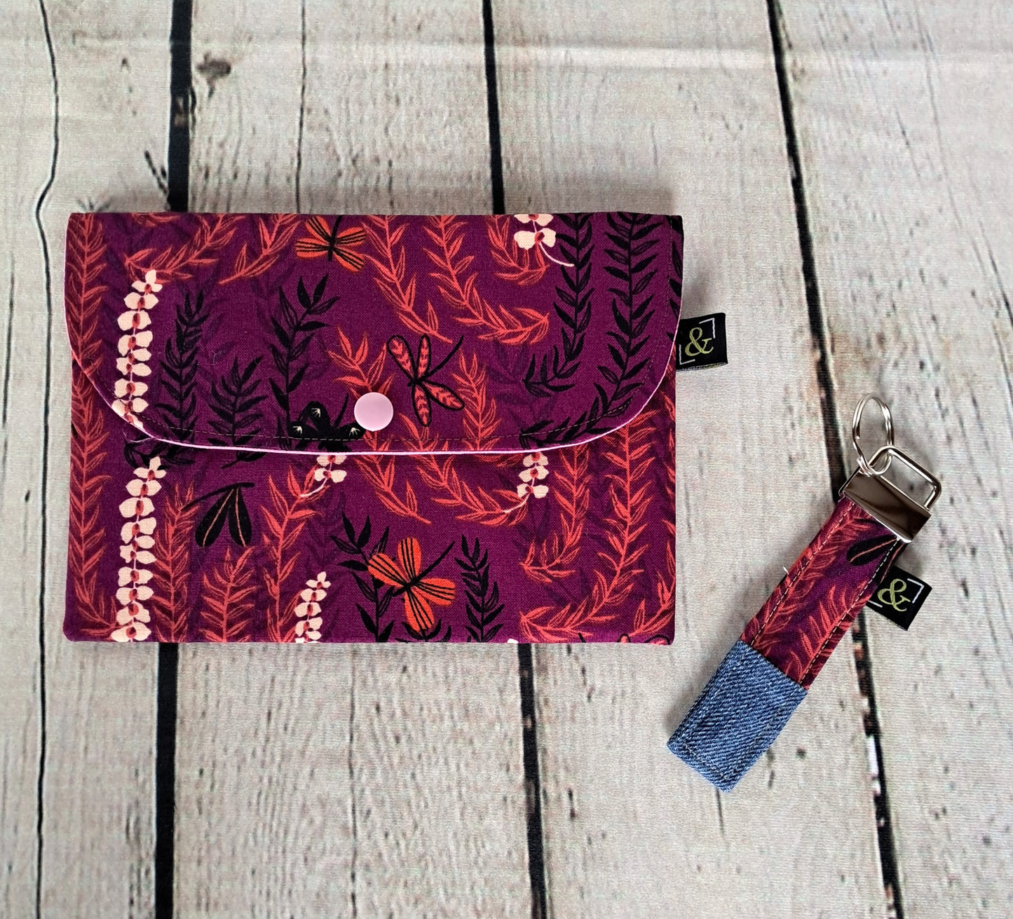 Conjunto estuche y llavero set featuring a handmade purple floral clutch pouch and matching keychain on weathered white wooden background. Botanical print fabric with burgundy base and orange leaves, black stems, and delicate pink flower details. Denim accent on keychain.