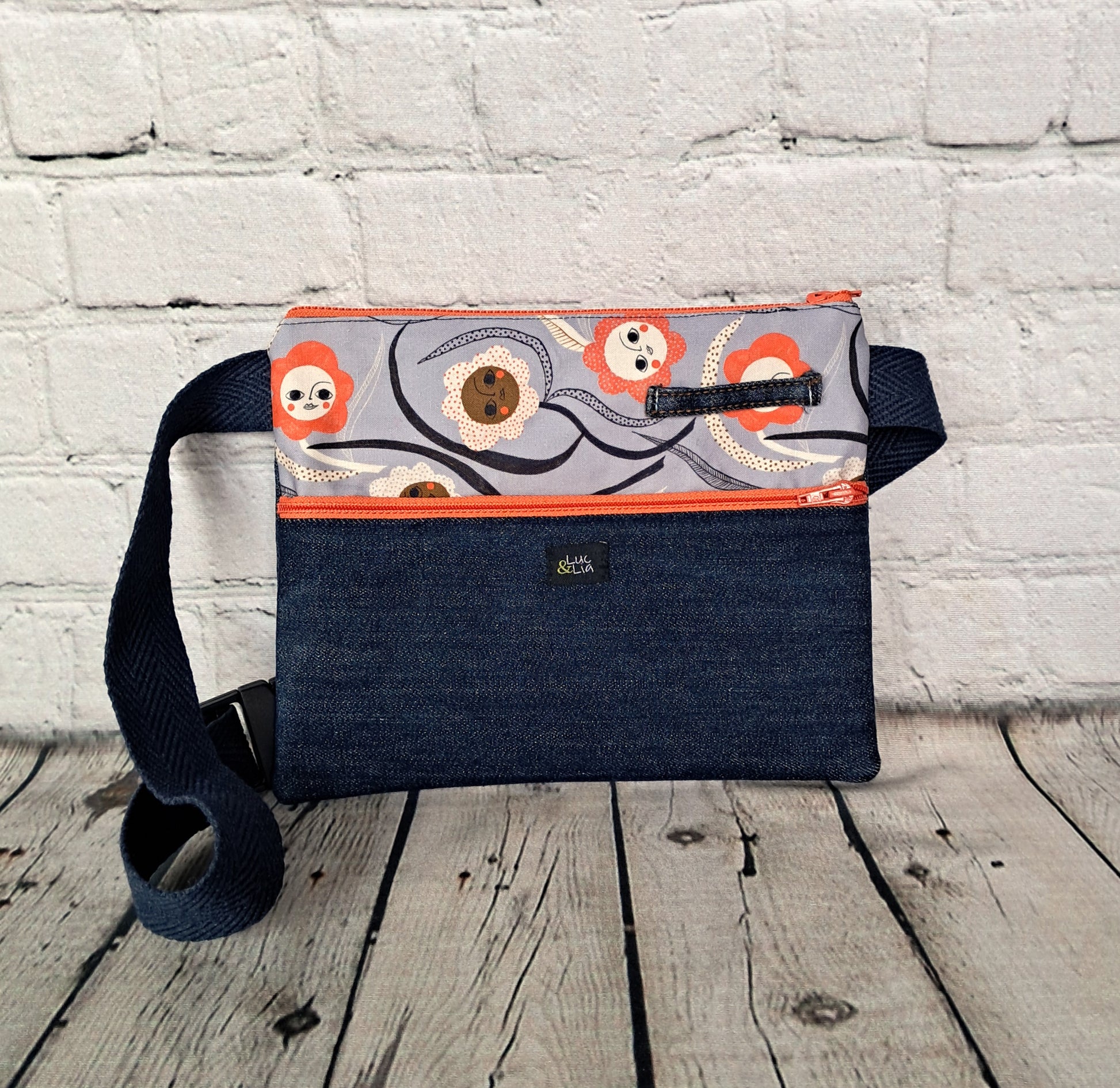 Riñonera diseño waist bag featuring a unique pattern with orange circular motifs on gray fabric, combined with dark blue denim. Handcrafted fanny pack with adjustable navy strap, orange zipper detail, and compact size perfect for urban style. White brick wall background showcases sustainable fashion accessory.