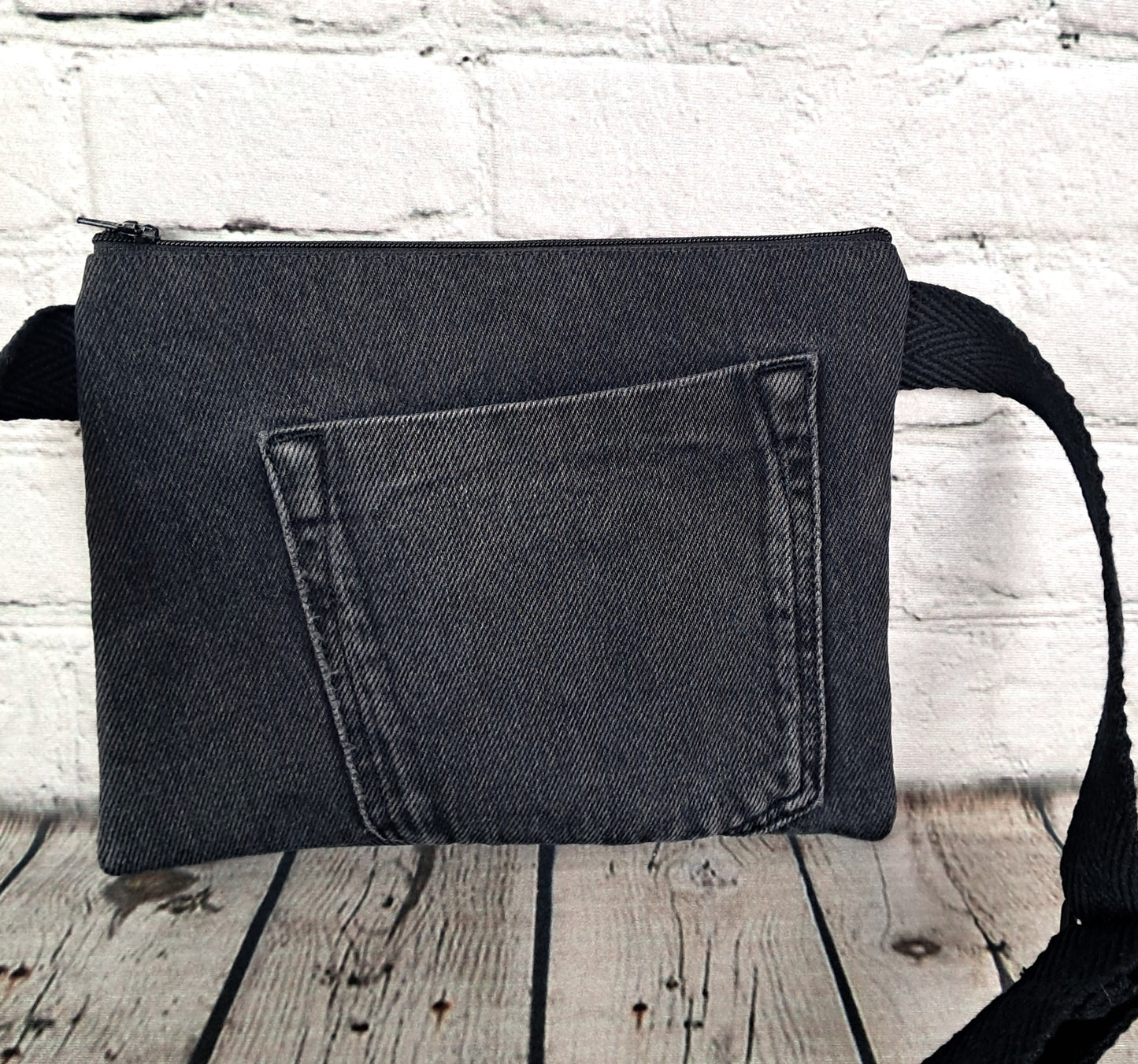 Rinonera diseno rojo y denim negro fanny pack featuring black denim fabric with pocket detail, adjustable black strap, against white brick wall background. Handcrafted sustainable accessory measuring 22 x 17 cm.