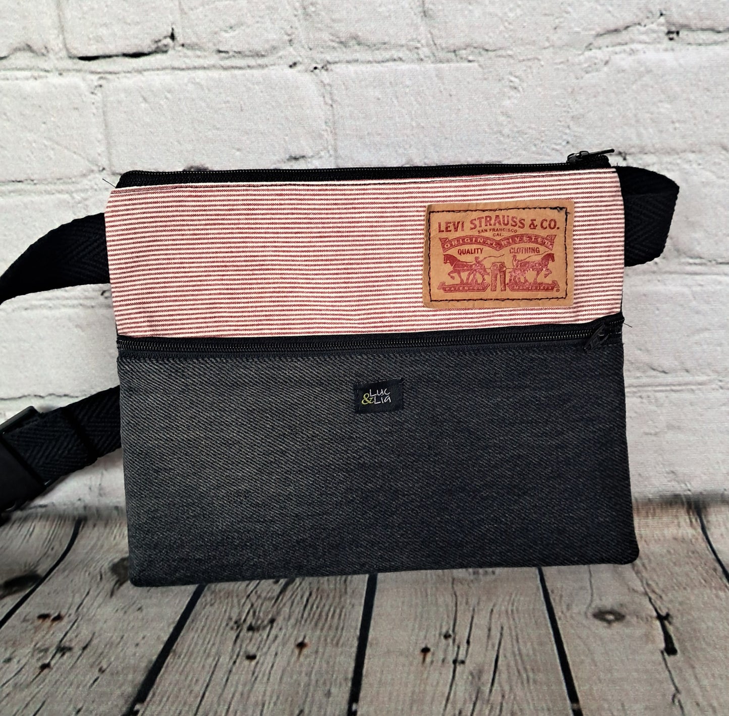 Riñonera diseño rojo y denim negro: Handcrafted fanny pack featuring red and white striped fabric with Levi's leather patch detail and black denim bottom. Sustainable upcycled design against white brick wall background. Adjustable black strap and zipper closure.