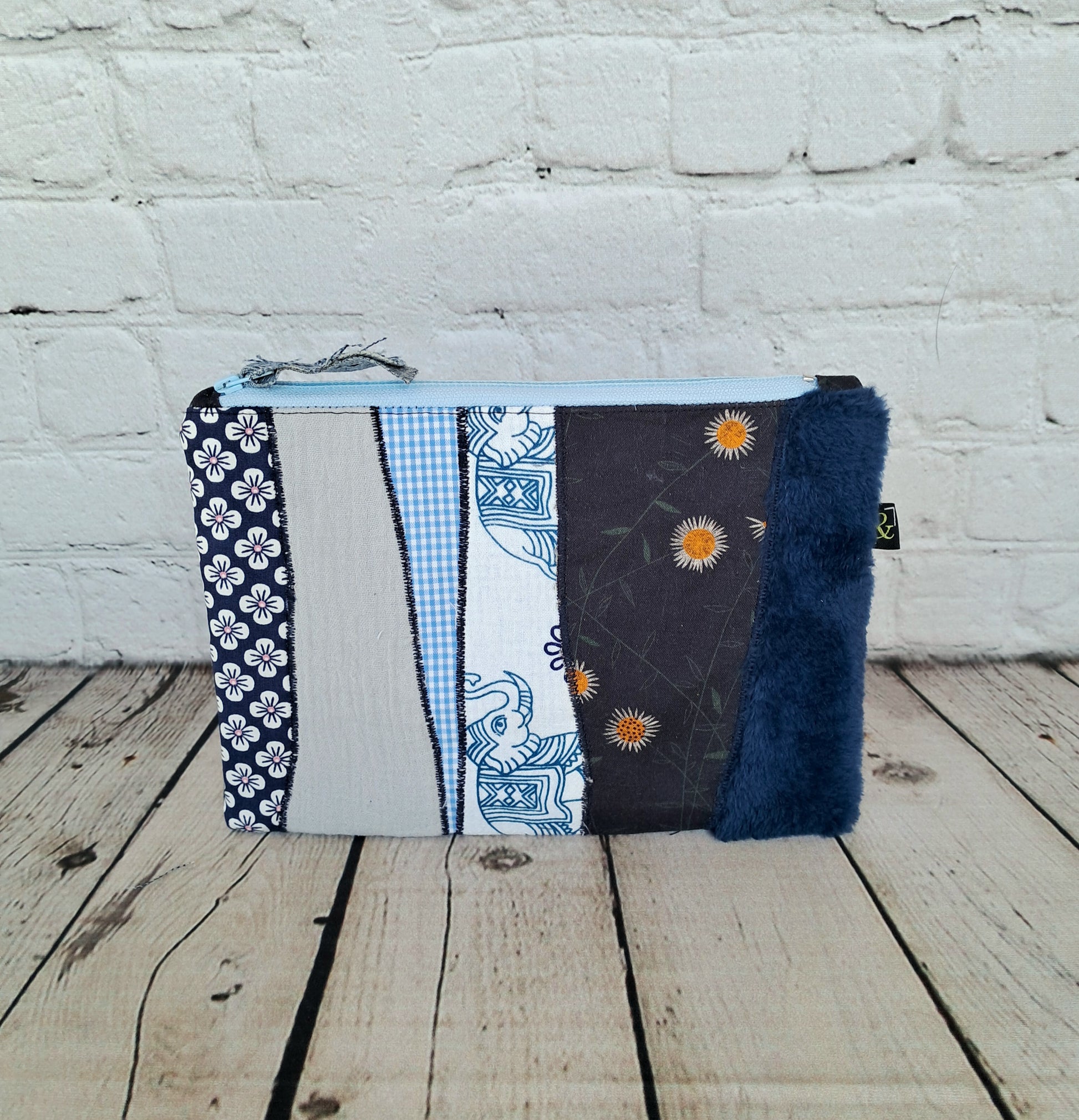 Estuche cremallera de retales azules: Handmade zipper pouch featuring patchwork design in blue tones with mixed patterns including gingham, floral prints, and faux fur. The case showcases various fabric textures against a white brick background, displaying sustainable craftsmanship and unique aesthetics.