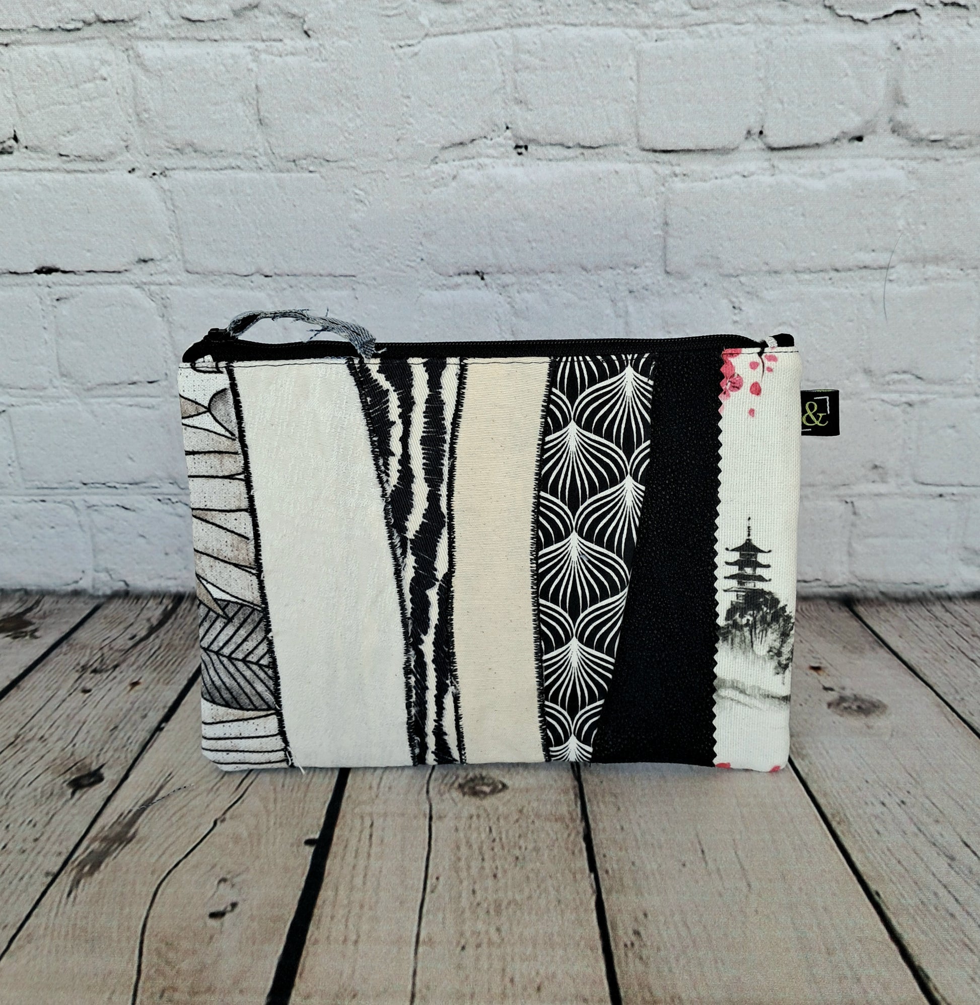 Estuche cremallera de retales blanco y negro - Handcrafted zipper pouch featuring patchwork design in black and white patterns, including geometric shapes, palm leaves, and Asian-inspired motifs on a rustic wooden surface against white brick wall background. Size 22 x 16 cm.