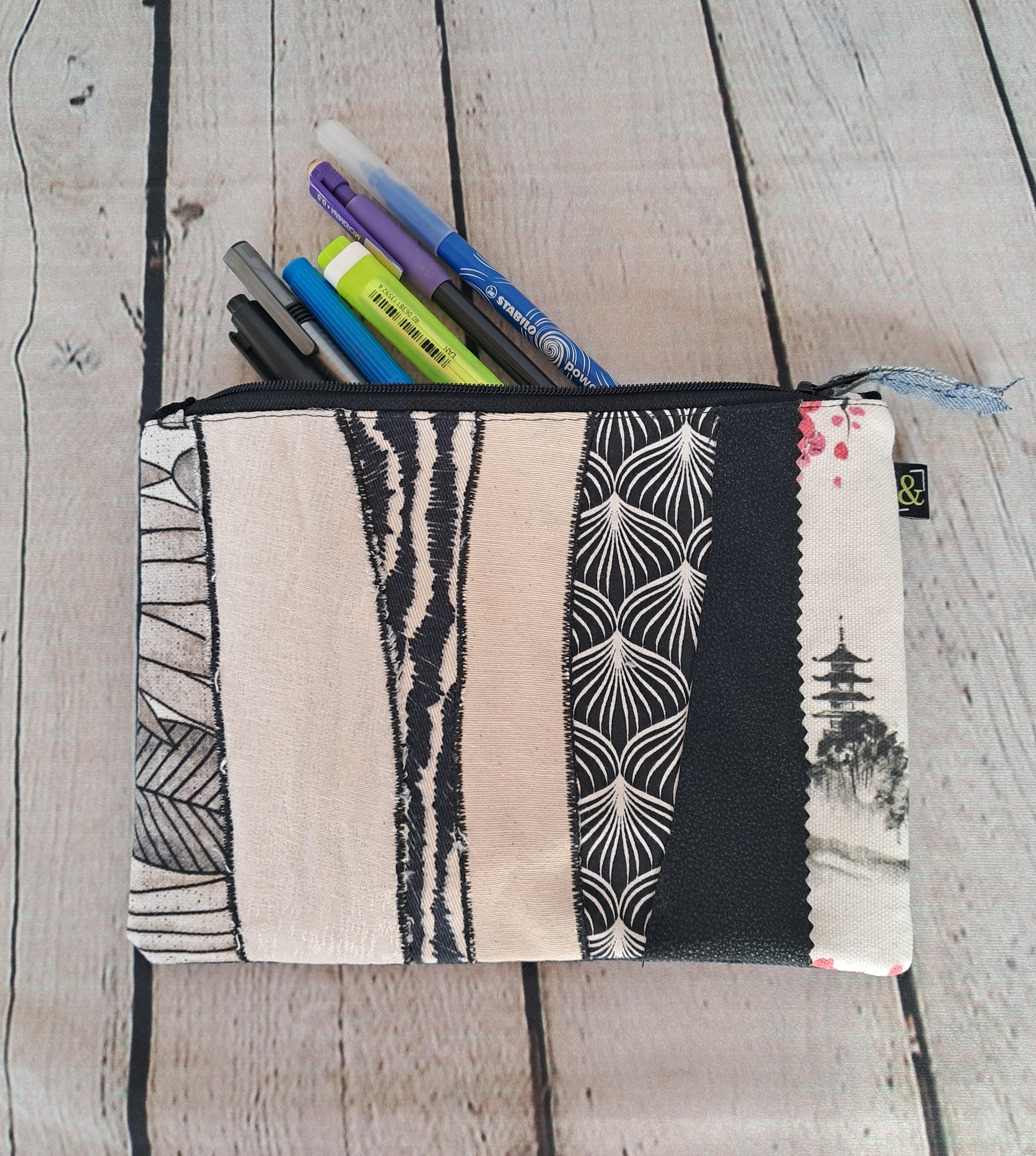 Estuche cremallera de retales blanco y negro: Handmade pencil case with black and white patchwork fabric design, featuring various patterns including geometric shapes, palm leaves, and Asian-inspired motifs. Shown partially open with pens inside, displayed on whitewashed wooden surface.