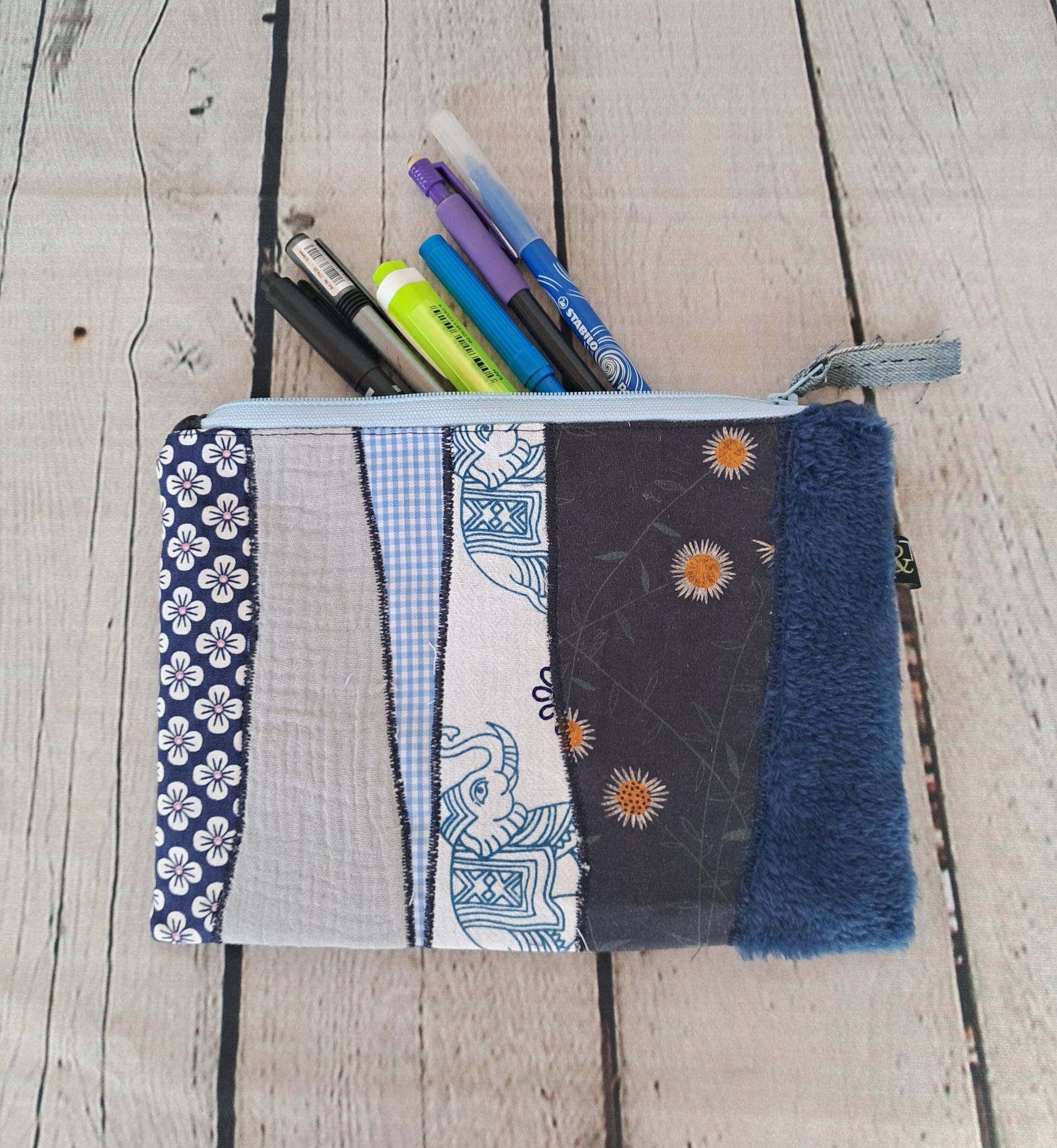 Handmade Blue Patchwork Pencil Case 'Estuche cremallera de retales azules' featuring mixed fabric patterns including floral prints, gingham, and textured materials, with blue zipper closure, shown with markers inside on wooden background. Size 22 x 16 cm.